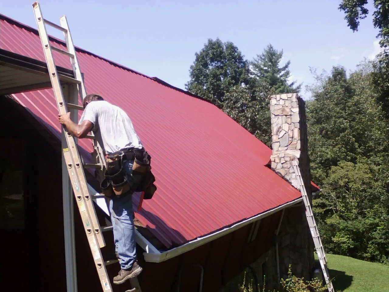 Roofer on a ladder beside a red metal roof and stone chimney — Soffit & Fascia Repair by Roofing Brothers in Blachly